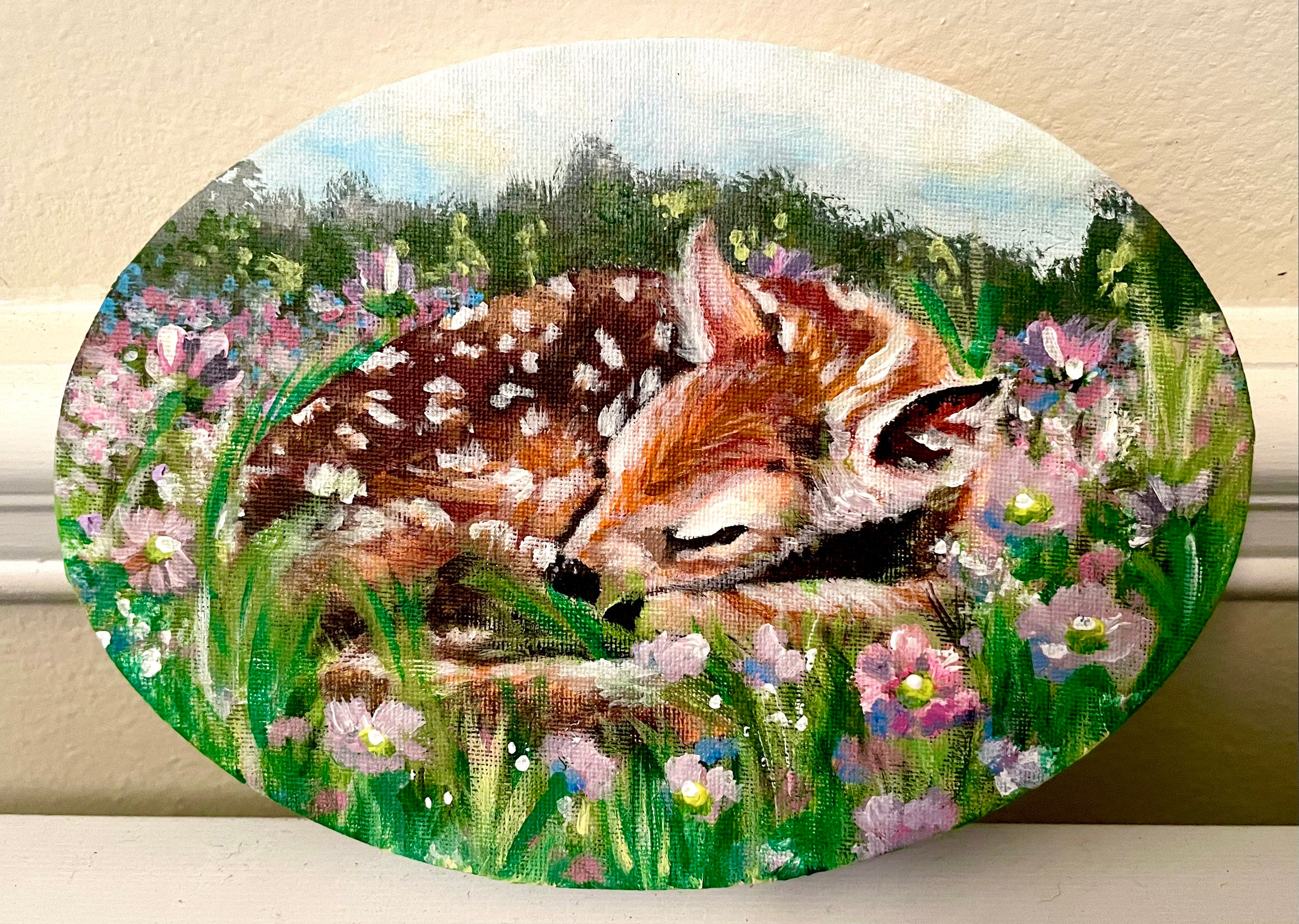 A sleeping fawn nestles among green grass and pink and purple wildflowers. A quiet moment, held in an oval canvas.